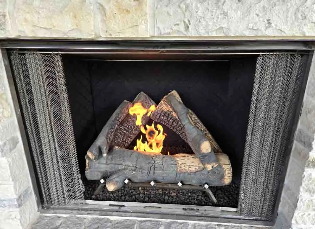 Close-up of Courtyard 42-inch outdoor gas fireplace Close-up of Courtyard 42-inch outdoor gas fireplace with black herringbone interior panels, standard definition gas logs burning with open flames, chain mesh screen, and natural stone surround.