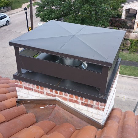 Rooftop close-up of a custom decorative chimney upper cap with dark metal pyramid roof panel. Rooftop close-up of a custom decorative chimney upper cap with dark metal pyramid roof panel and open sides, mounted on a white and red brick chimney with clay tile roof visible below.