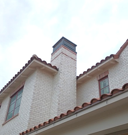 Custom decorative chimney upper atop a full white brick chimney. Custom decorative chimney upper atop a full white brick chimney with red brick detailing on a Spanish-style home with clay tile roof and dormers.