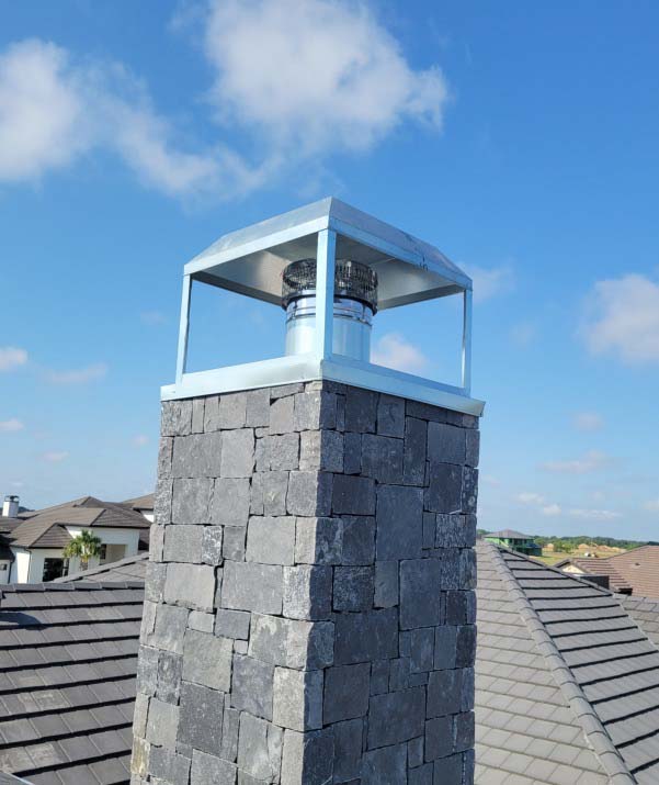 Decorative Upper model DEC-1 in standard galvanized metal mounted on a dark stone chimney. Decorative Upper model DEC-1 in standard galvanized metal mounted on a dark stone chimney, viewed from rooftop level with clay tile roof and suburban neighborhood in the background.