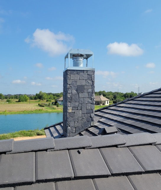 Decorative Upper model DEC-1 in standard galvanized metal installed on a tall dark stone chimney. Decorative Upper model DEC-1 in standard galvanized metal installed on a tall dark stone chimney with gray tile roof in the foreground and a scenic waterfront and green landscape in the background.