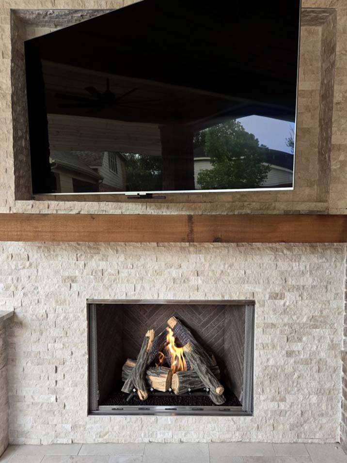 Courtyard 42-inch outdoor gas fireplace set in a stacked stone wall. Courtyard 42-inch outdoor gas fireplace with brown herringbone panels set in a stacked stone wall below a wood mantel and TV on a covered patio in Cypress, TX.