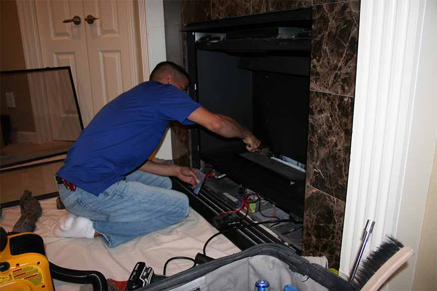 Perfection Supply technician servicing the burner area of a gas fireplace with the log set and glass front removed in Houston, TX.